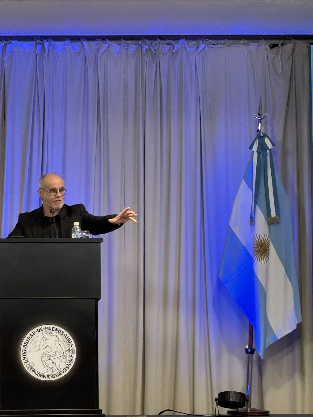 Carlos Moreno delivering the inaugural lecture at a lectern bearing the seal of the Universidad de Buenos Aires, beside the Argentine flag