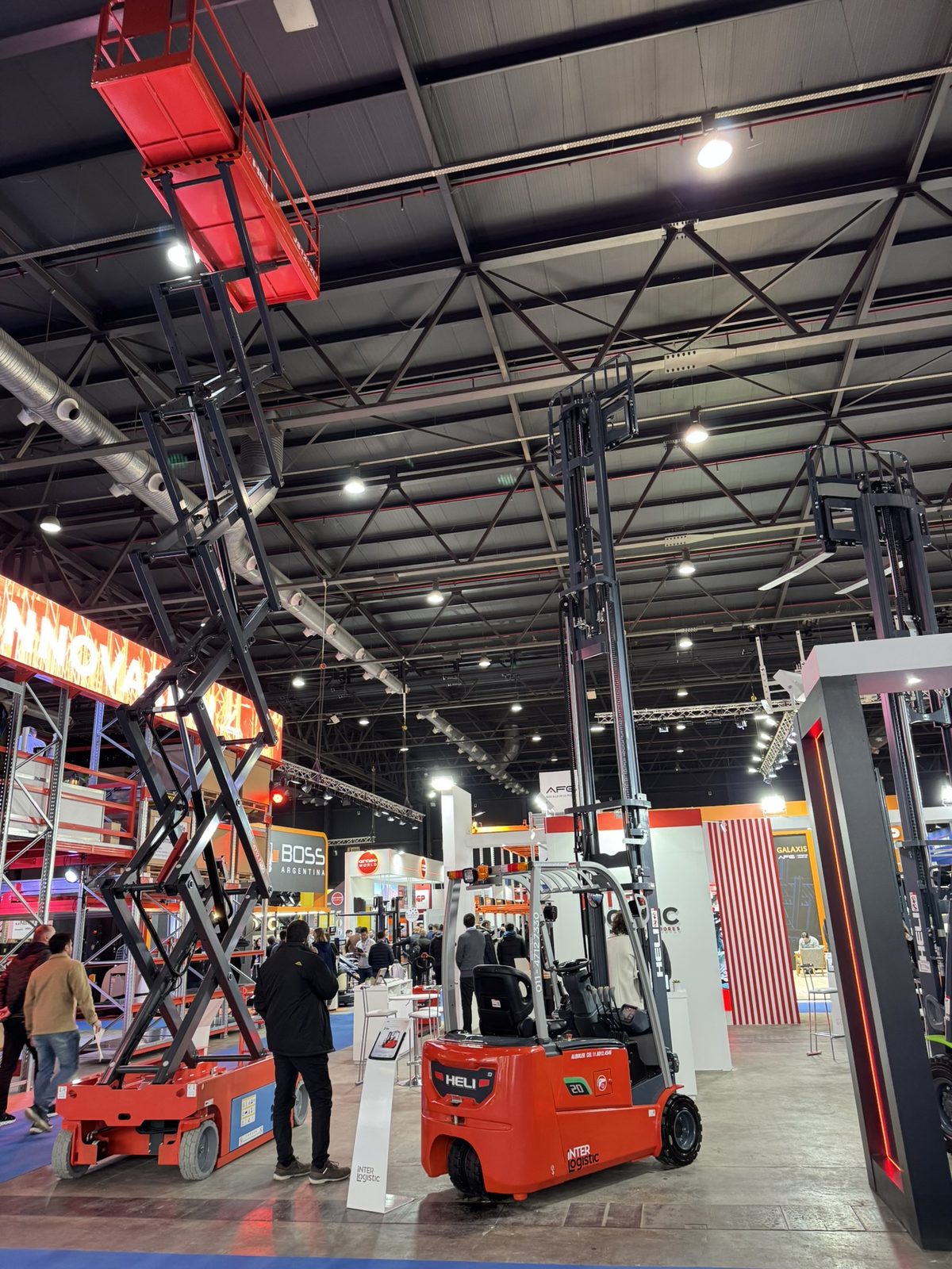 Exhibition floor at Expo Logisti-k 2026 in La Rural, Buenos Aires, with a scissor lift extended to full height, a high-mast electric forklift and pallet racking under the pavilion roof