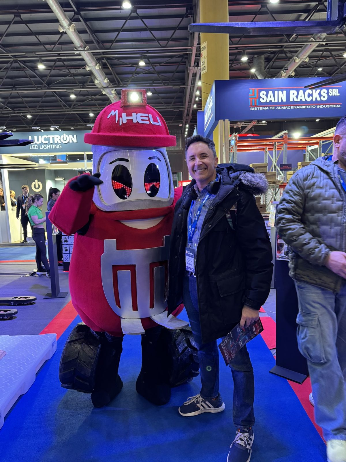 Sergio Mastrogiovanni of Socradata with the Heli forklift mascot on the exhibition floor at Expo Logisti-k 2026, with the Sain Racks and Luctron stands behind