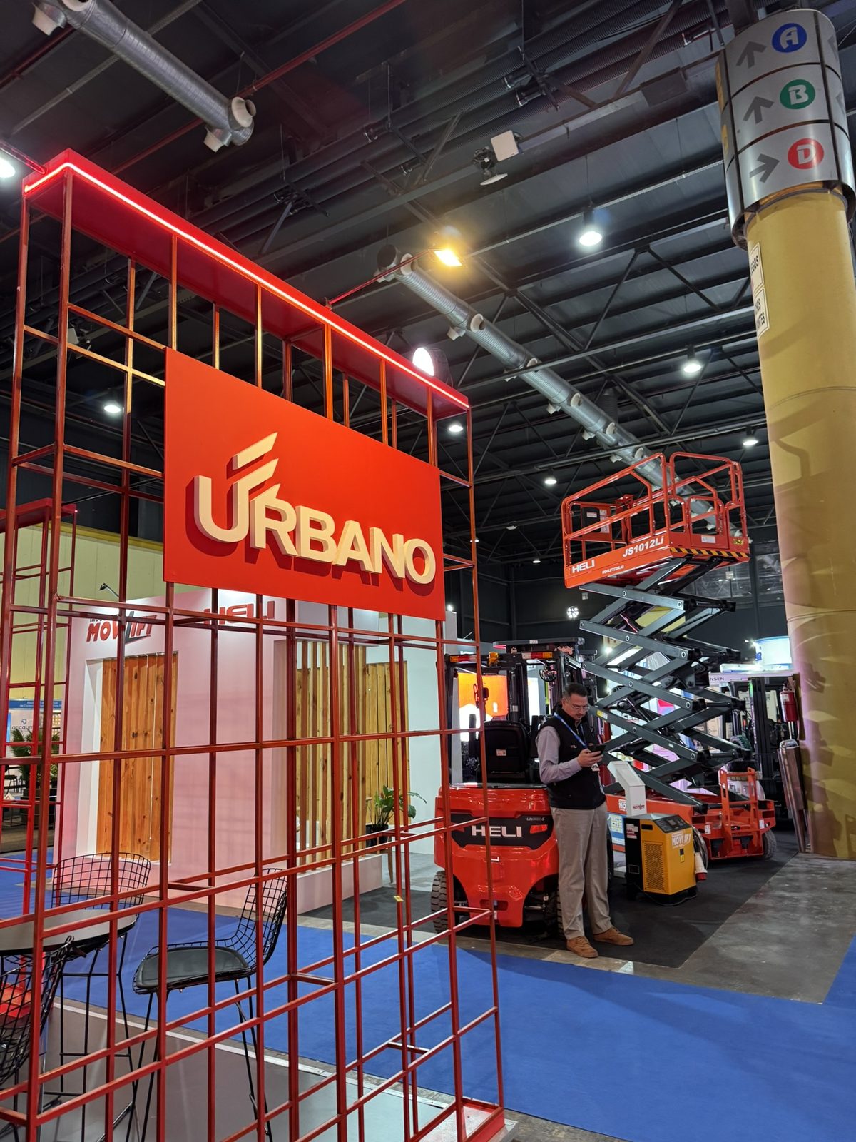 Stand at Expo Logisti-k 2026 with a Heli electric forklift, a JS1012Li lithium scissor lift and a battery charging unit, beside the Urbano and Movilift stands