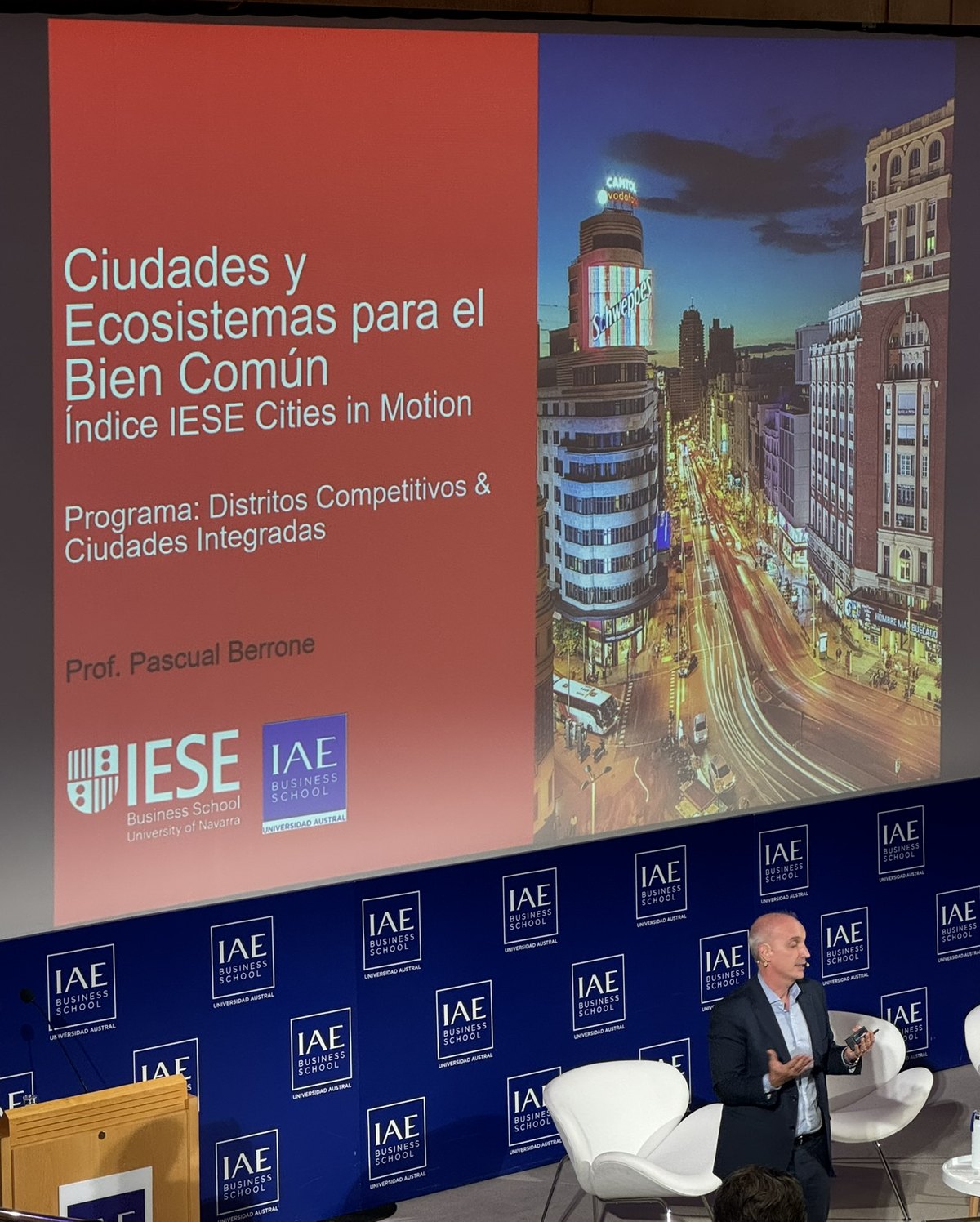 Pascual Berrone of IESE speaking beside a projected title slide reading Ciudades y Ecosistemas para el Bien Comun, Indice IESE Cities in Motion, Programa Distritos Competitivos y Ciudades Integradas, against an IAE Business School backdrop